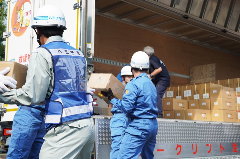「八王子市災害」と書かれた白いヘルメットと「八王子市」のビブスを着用した職員たちが、熊本県八代市へ送る支援物資の段ボール箱を救援物資輸送トラックの荷台へ次々と積み込む様子