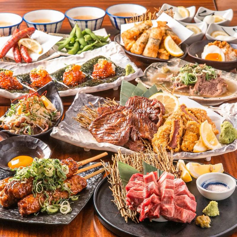 木のテーブルに並ぶ肉料理の盛り合わせ。中央の大皿には焼き目の付いた厚切り牛タンと断面が赤い牛カツ、レモンとわさび。手前左につくねと生卵、手前右に黒皿のサイコロ状の生肉、左にイクラとウニの海苔巻き、奥に枝豆やソーセージ