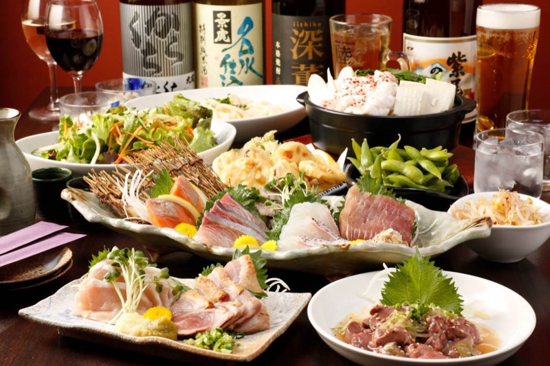 木のテーブルいっぱいに並ぶ宴会料理。中央に葉形の皿の刺身盛り合わせ、手前に炙り肉とレバー、左にサラダ、奥に天ぷらと黒い鍋、枝豆、焼酎の一升瓶、ビールジョッキとワイングラス