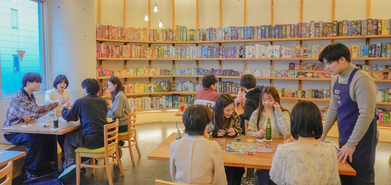 壁一面の棚にボードゲームが並ぶJELLY JELLY CAFEの店内。木のテーブルで複数の客がゲームをし、右側にはJELLY JELLY CAFEと書かれたエプロンのスタッフが接客している