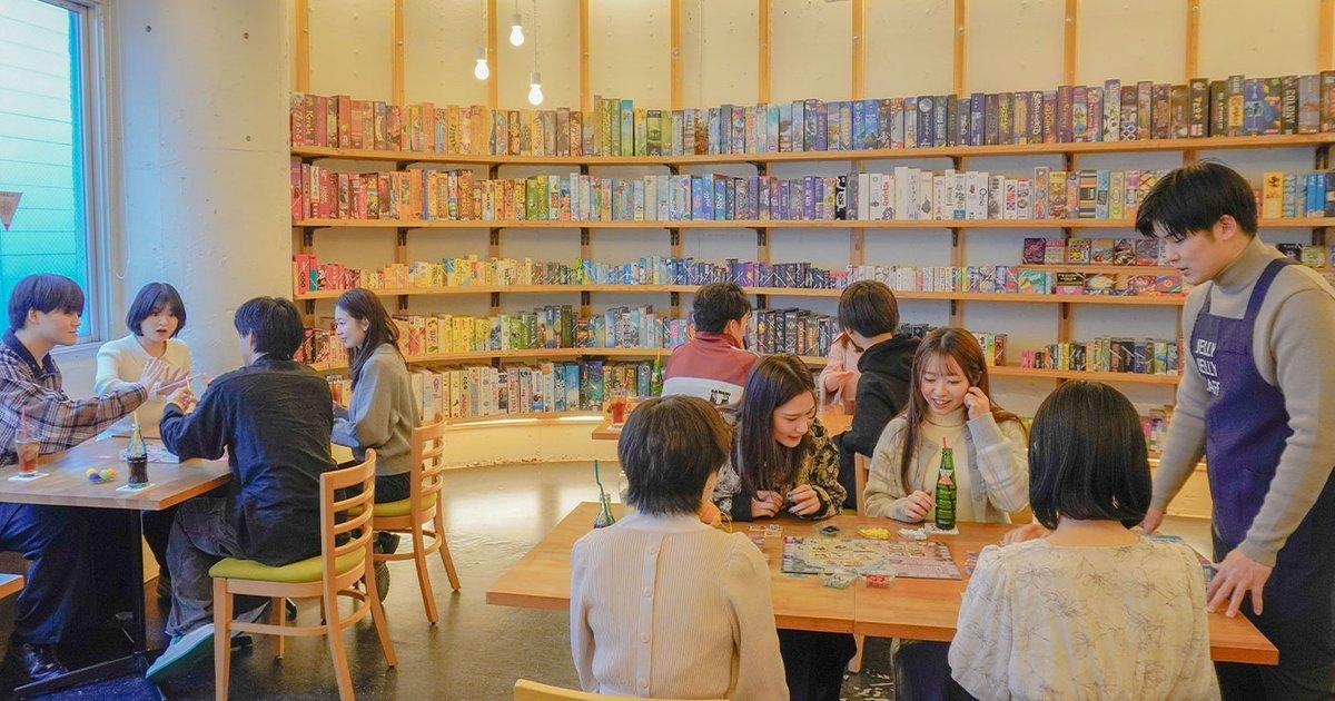 壁一面の棚にボードゲームが並ぶJELLY JELLY CAFEの店内。木のテーブルで複数の客がゲームをし、右側にはJELLY JELLY CAFEと書かれたエプロンのスタッフが接客している