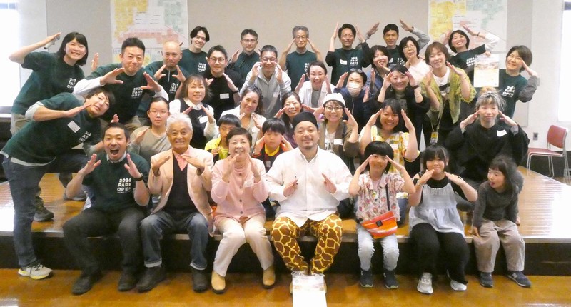 室内で円を組む参加者たち。中央の床に桑都の杜のロゴとそうとうたのしい桑都の杜の文字が描かれた布が広がり、周囲の椅子に座る大人と子どもが布を囲んでいる