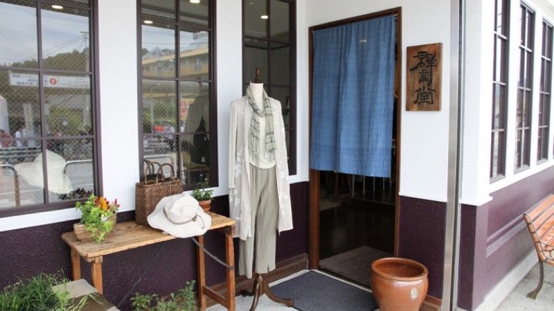 白い壁と暗い紫色の腰壁の店舗入口。右側に群言堂と縦書きされた木札、入口上部に藍色ののれん、左側にカーディガンを着たマネキンと帽子や籠を置いた木テーブルがある