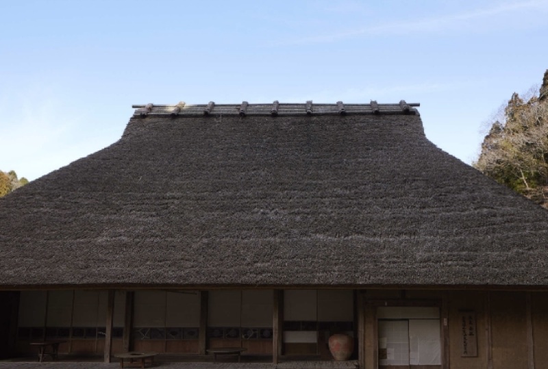 青空の下に、棟を丸太で押さえた大きな茅葺き屋根の建物があり、軒下に障子と縁側が見える