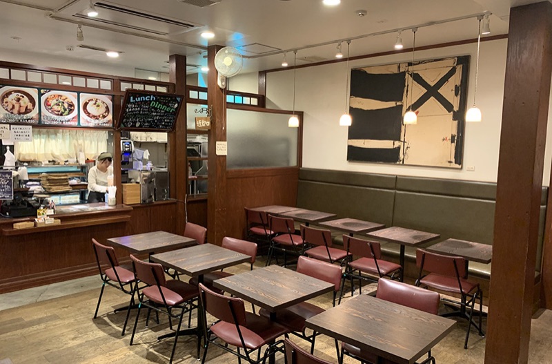 木の柱とテーブルが並ぶカフェ店内。左のカウンター上にカレーなどのメニュー写真とLunch & Dinnerの黒板、右の壁に大きな抽象画と緑のベンチ席がある