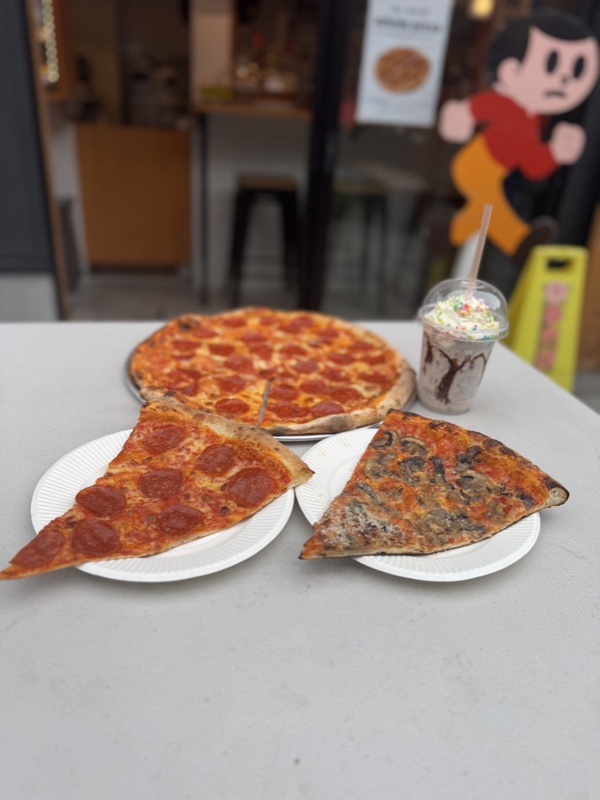屋外のテーブルにペパロニとキノコの大きなスライス2枚、奥にペパロニのホールピザとホイップクリーム付きのシェイクが並び、背景に店舗入口がぼやけて見える