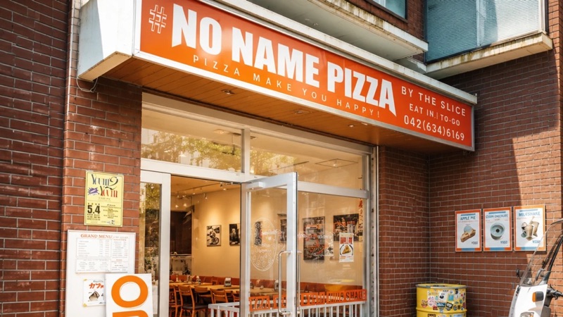 赤いレンガ壁の店舗入口。上部に#NO NAME PIZZAとBY THE SLICEと書かれたオレンジの看板、ガラスドアの脇にメニュー掲示と黄色いドラム缶がある