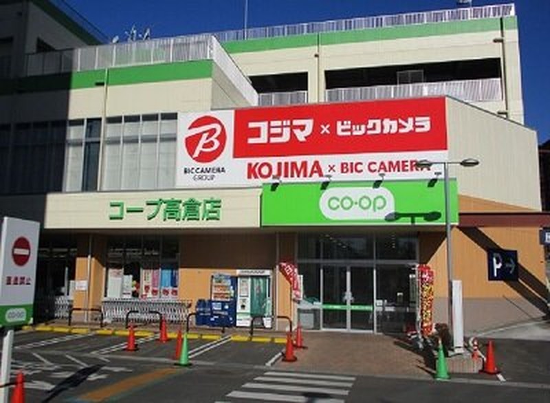 白い外壁の商業施設。1階左に緑地のコープ高倉店看板、中央に赤いコジマ×ビックカメラの大型看板、右のガラス扉上に緑のco-opロゴ。手前の駐車スペースにカラーコーンが置かれている