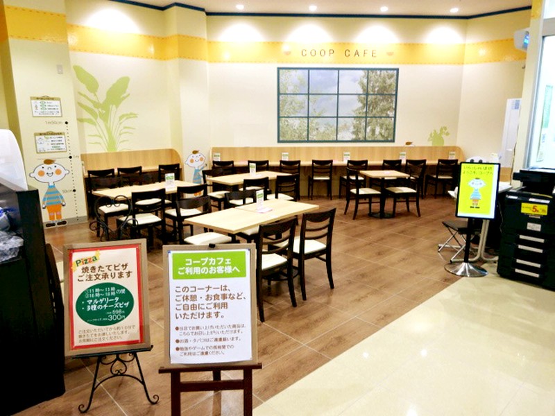 COOP CAFEと書かれたイートイン。木目の床に白いテーブルと椅子、奥の壁に緑のイラスト、手前に焼きたてピザの案内と利用ルールの看板が置かれている