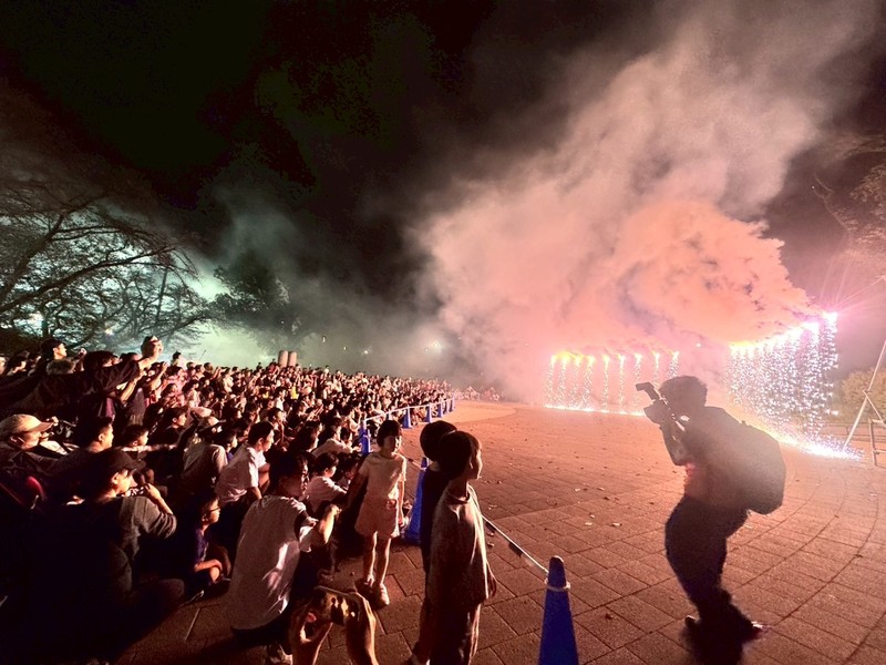 夜の広場で青いロープの内側に並ぶ仕掛け花火から白い火花が落ち、ピンクとオレンジの煙が上がる。左側には家族連れの観客、手前右にはカメラを構える人の影がある