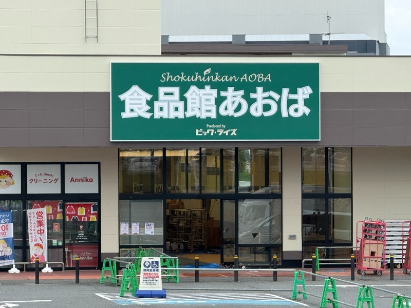 緑の看板に白い文字で食品館あおば、上にShokuhinkan AOBA、下にProduced by ビッグ・ライズ。ガラス扉の入口は養生中で、左隣にクリーニングAnnika、手前に緑のカラーコーンと車椅子マークの看板