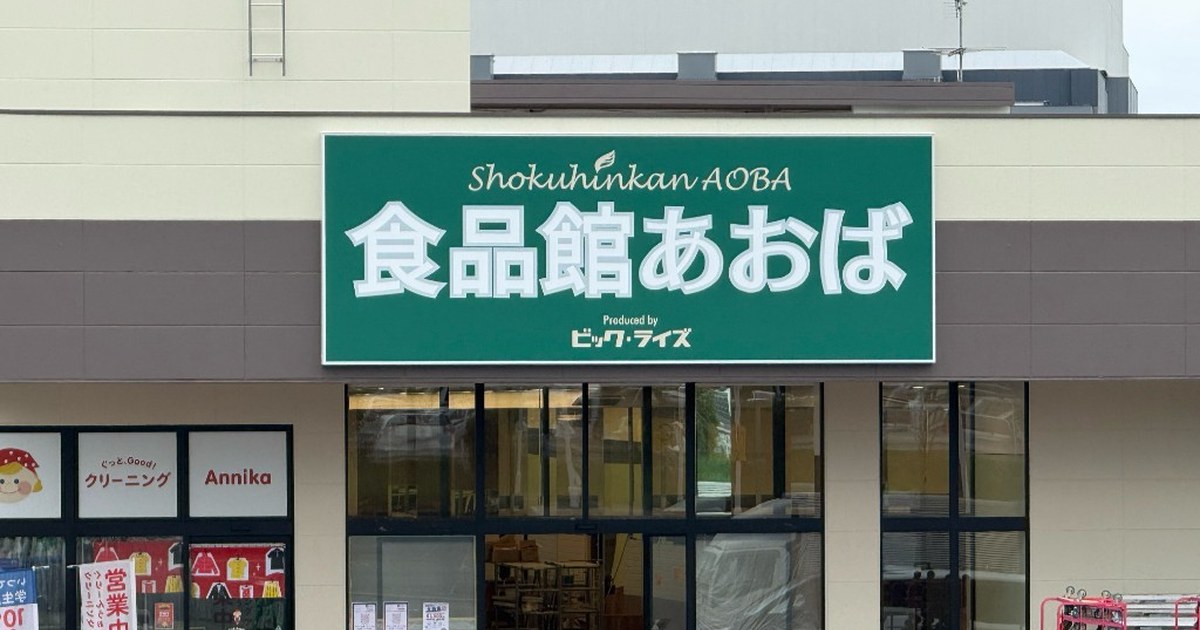 緑の看板に白い文字で食品館あおば、上にShokuhinkan AOBA、下にProduced by ビッグ・ライズ。ガラス扉の入口は養生中