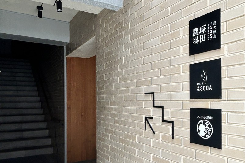 白いタイル壁の右手に塚田農場、BAR&SODA、八王子焼肉の黒い案内板が縦に並び、中央に階段を示す黒い線、左手に暗い階段がある