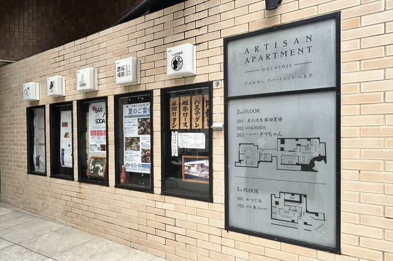 白いタイル壁の窓にARTISAN APARTMENT HACHIOJIの案内図。2階203に八王子焼肉タマちゃん、201に塚田農場、1階に鮨つぐみと焼鳥卓と記されている