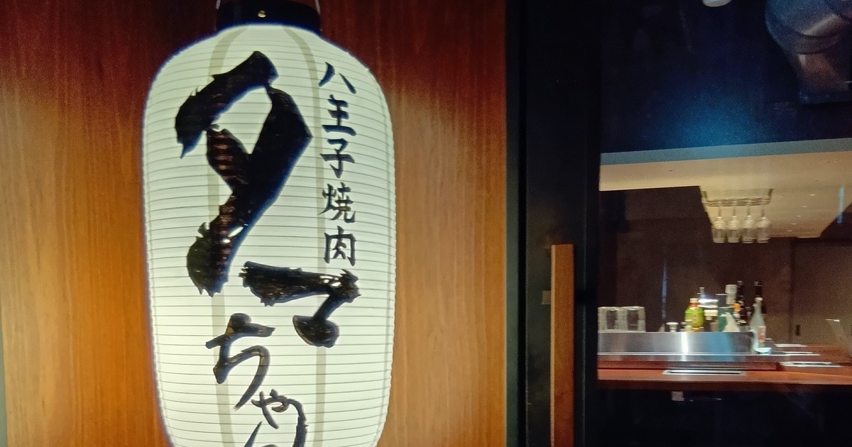 木の壁に白い提灯が掛かり、筆文字で八王子焼肉とタマちゃんと書かれている。右奥は暗いカウンター席