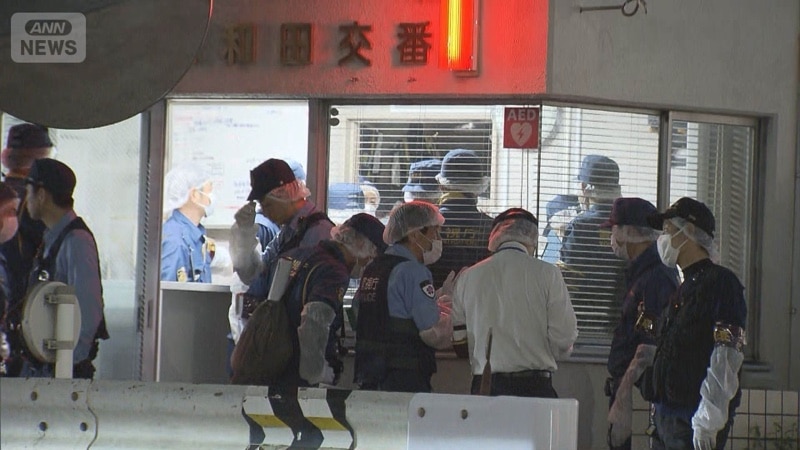 夜間の大和田交番入口。白い壁に赤い縦帯と大和田交番の表示があり、マスクをした警察官らがガラス戸の前に集まっている