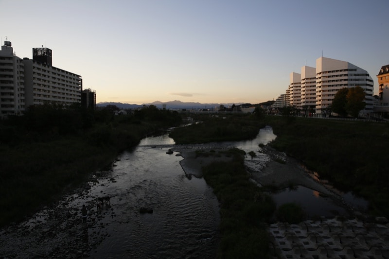 夕暮れの大和田橋から見た浅川。川面の向こうにマンションと白い建物が並び、遠景に山並みが見える