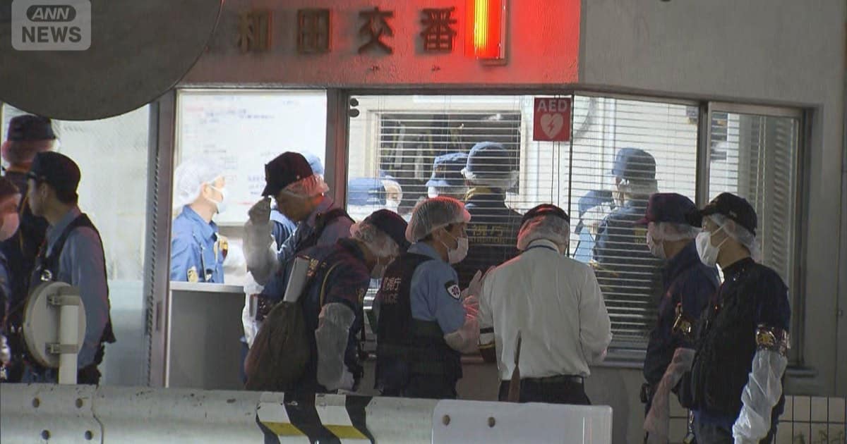 夜間の大和田交番入口。白い壁に大和田交番の表示があり、マスクをした警察官らがガラス戸の前に集まっている