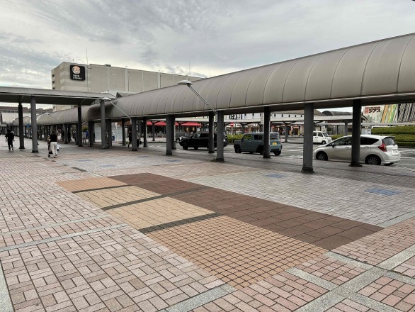 南大沢駅前の屋根付き歩行空間。ベージュと茶色の舗装見本が床に敷かれ、奥に柱の並ぶ通路と道路を走る車、白い建物が見える