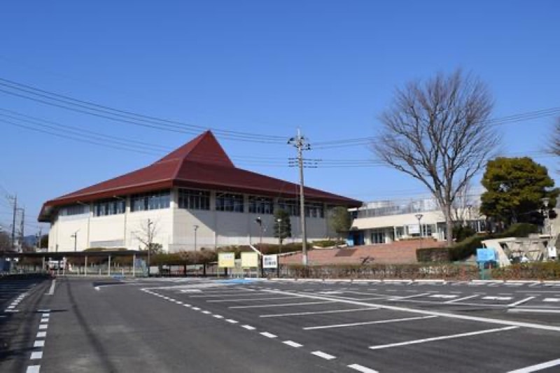 青空の下に建つ赤い三角屋根のクリーム色の体育館。手前は白い区画線の駐車場で、右に葉の落ちた木と電柱がある