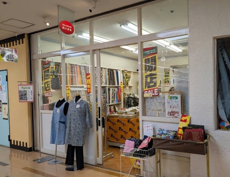 ガラス戸の店内に生地の棚とエプロン姿のマネキン。赤い看板にめいぷるとあり、黄色い垂れ幕に閉店とSALEと書かれている