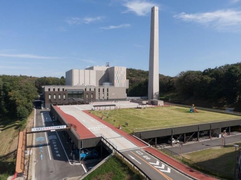 館クリーンセンターの全景。高い煙突と工場棟、屋上に芝生の広場、手前に搬入路と車両の入口が見える
