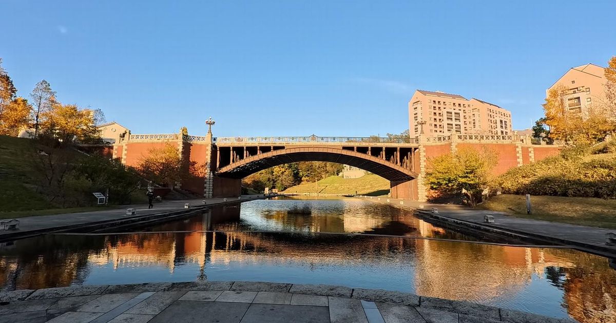 水面に映る赤レンガのアーチ橋。欄干に装飾があり、奥に丘と建物が見える