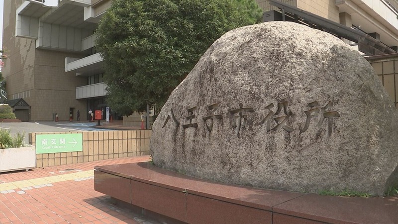 八王子市役所と刻まれた大きな石。奥に市役所の建物と木、左に南玄関の案内板がある