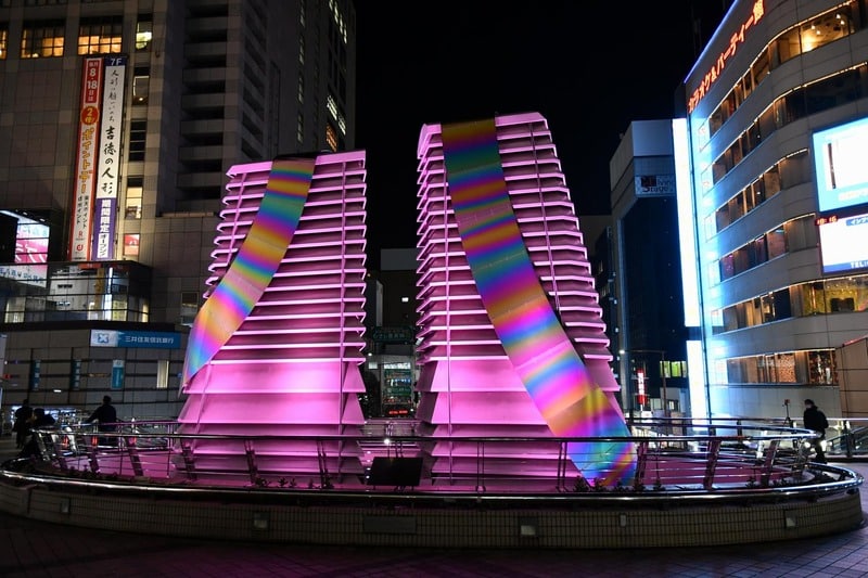 夜の駅前広場で、二基の塔がピンクと虹色に照らされている。手前は円形の柵で囲まれ、奥にビルの明かりが見える