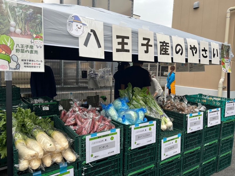 白いテントの下に八王子産のやさいと書かれた看板。大根や葉物が入った緑のかごが並び、奥に人が立っている