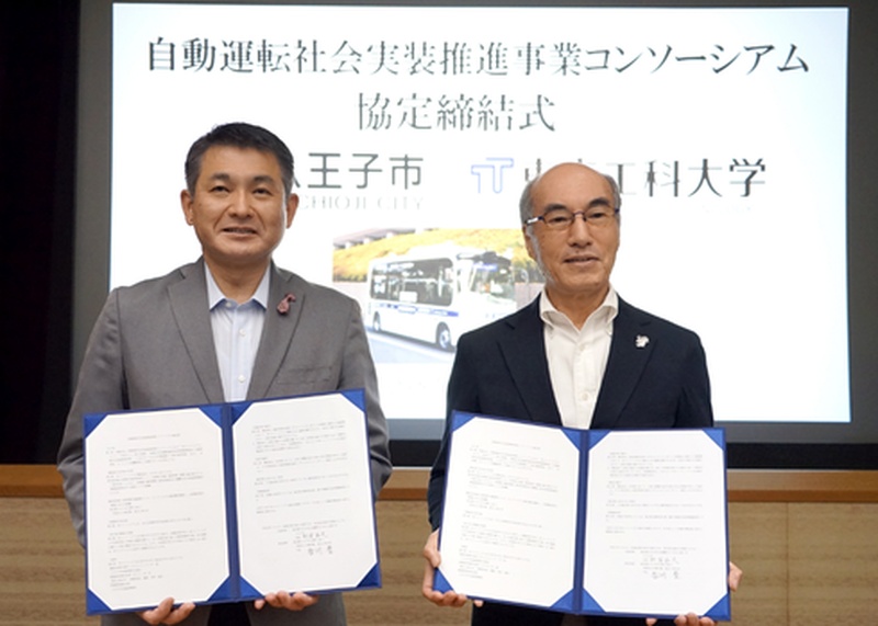 「自動運転社会実装推進事業コンソーシアム協定締結式 八王子市 東京工科大学」と映されたスクリーンの前で、協定書を開いて持つ八王子市長と東京工科大学学長