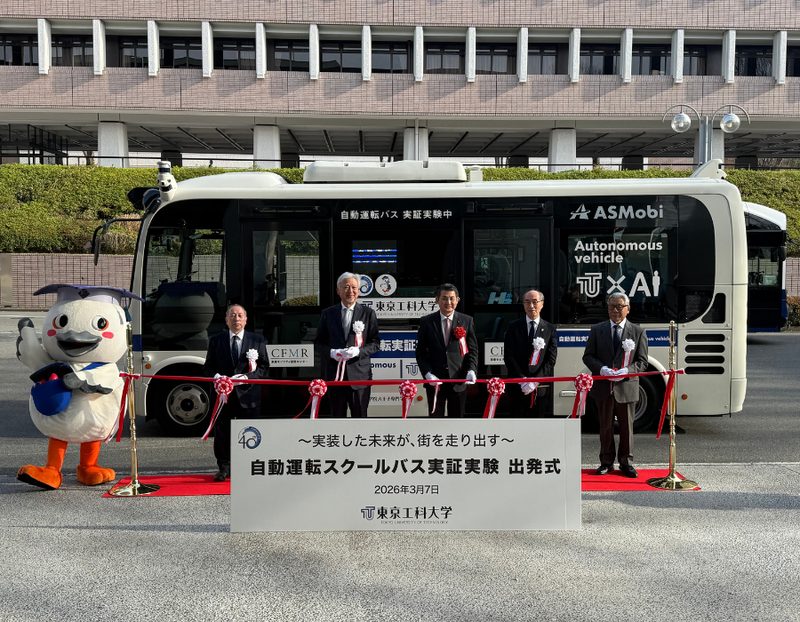 「自動運転スクールバス実証実験 出発式 2026年3月7日 東京工科大学」と書かれた看板の前で、自動運転バスとテープカットの紅白リボンを背にして並ぶ関係者5人とマスコットキャラクター