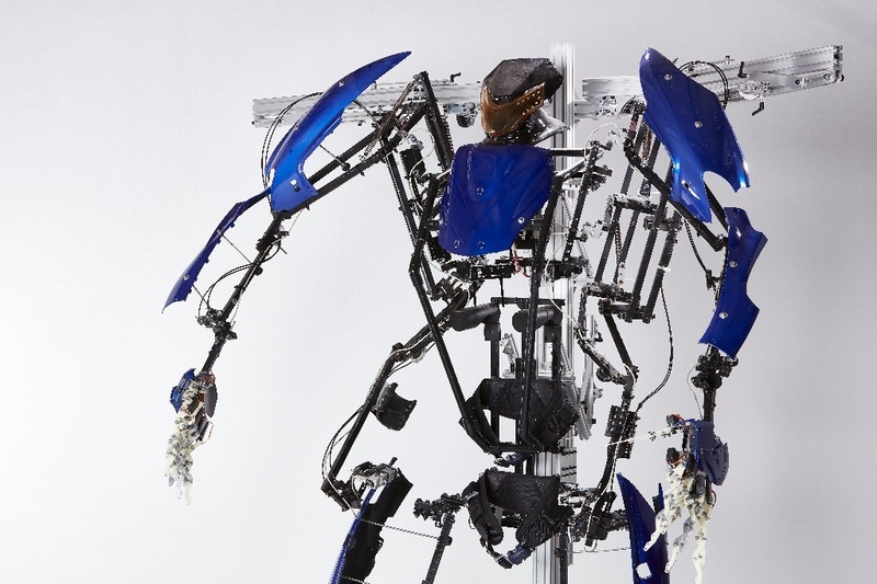 黒いフレームに青い外装パーツを組み合わせた人型の装着型ロボットの上半身。両腕は白い指を持つマニピュレーターにつながっている
