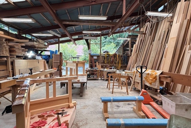 トタン屋根の木造作業場の内部。加工途中の木の枠組みや作業台が並び、奥の壁際には長い木材が立てて何枚も保管されている