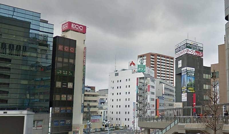 曇天下のJR八王子駅北口。左にECCの赤い屋上看板がある黒い高層ビル、中央に白い中層ビル、右にペデストリアンデッキが伸びている