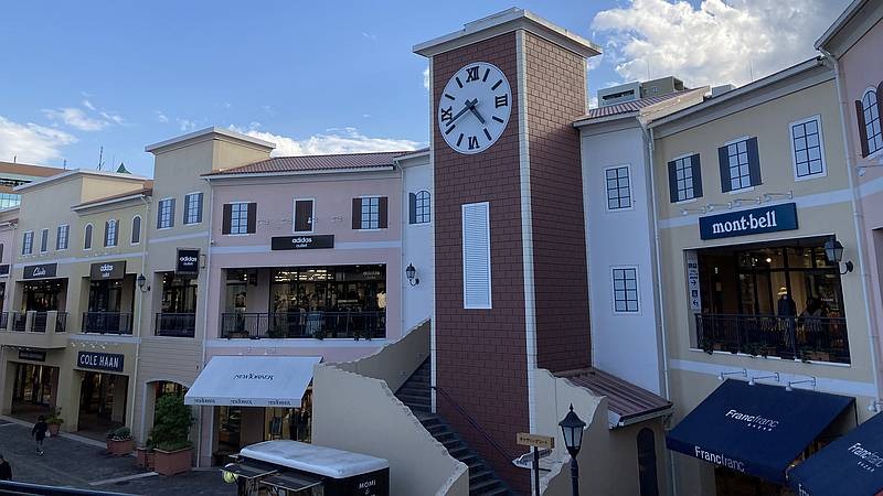 ベージュの店舗が並ぶアウトレットの広場。中央にローマ数字の白い時計が付いた茶色の塔、右にmont-bellとFrancfrancの看板