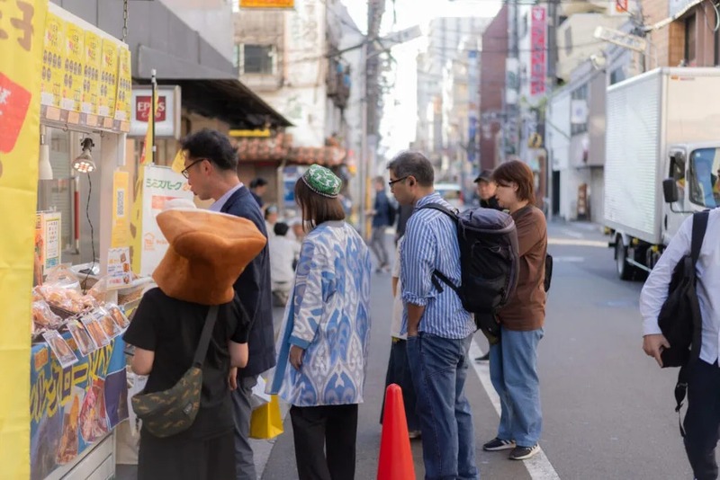 歩道の屋台前に並ぶ客。左端の人は茶色いパン型の帽子をかぶり、白いショーケースの前に立っている