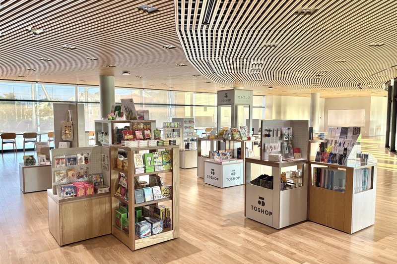 ガラス張りの明るい館内に、本や雑貨を並べた白と木目の陳列台が点在するTOSHOP 桑都の杜の売り場。手前の台にTOSHOPのロゴがあり、波打つ木製ルーバーの天井と奥の窓際に椅子が見える