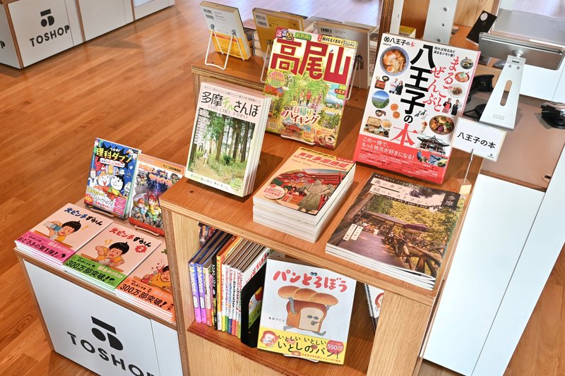 八王子の本と書かれた札が立つ木製の陳列台。高尾山のハイキング本、八王子の本と題したグルメ本、多摩の散歩本、パンどろぼうの絵本などが平積みや面陳列で並び、奥にTOSHOPのロゴ入りの台がある