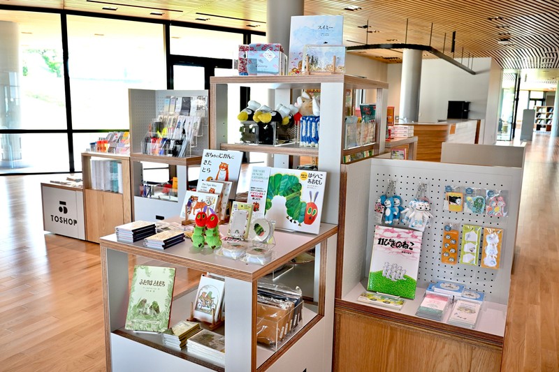 窓際の売り場に並ぶ絵本キャラクターグッズ。白い棚と木の台に絵本、緑色の人形、ぬいぐるみ、ポストカードが置かれ、右側の白い有孔ボードにキーホルダーや小物が吊るされている