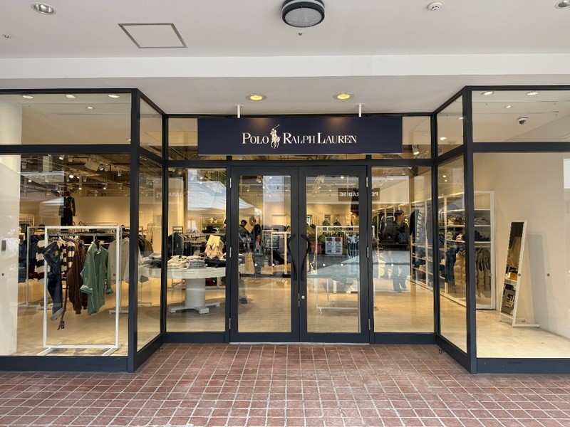ガラス張りのポロ ラルフ ローレン ファクトリーストアの正面入口。中央にネイビーのPOLO RALPH LAURENの看板があり、両開きのガラス扉の奥に服が掛かったラックが見える。手前の床はれんが調のタイル