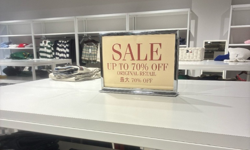 白い棚の上に置かれたセールの掲示。ベージュの台紙に赤い文字でSALE UP TO 70% OFF ORIGINAL RETAIL 最大70% OFFと書かれ、奥の棚にはチェックのシャツやボーダーのニットが並んでいる