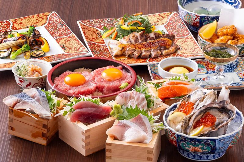木のテーブルに海鮮料理が並んだ宴会コースの写真。升に盛った刺身、卵黄をのせたユッケ丼、いくらをのせた生牡蠣、炙ったあなごの寿司、唐揚げなどが色鮮やかな器に盛られている