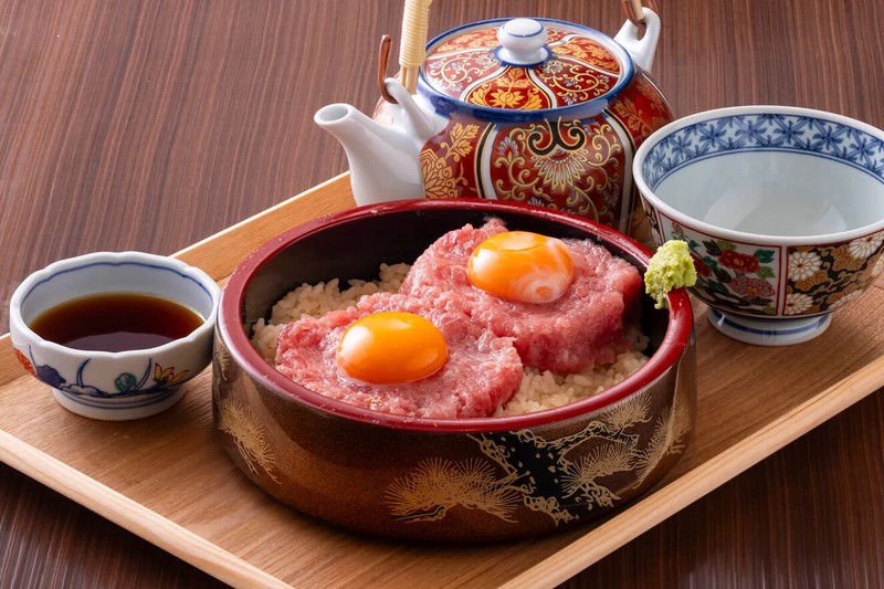 木の盆に置かれた赤い丼に、卵黄を2つのせたまぐろのユッケがご飯の上に盛られている。手前に醤油の小皿、奥に急須と湯のみがある