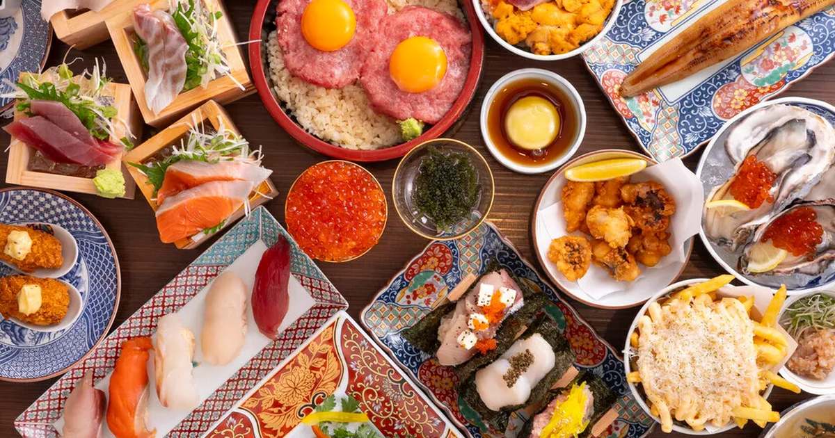 木のテーブルに海鮮料理が並んだ写真。卵黄をのせたまぐろのユッケ丼、升に盛った刺身、握り寿司、いくら、生牡蠣、手巻き寿司、唐揚げ、チーズをかけたポテトなどが色とりどりの器に盛られている