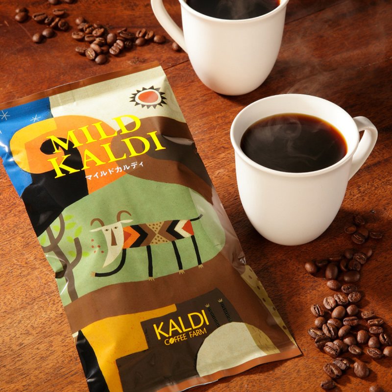 木のテーブルに置かれたカルディのコーヒー豆「MILD KALDI（マイルドカルディ）」の袋と、湯気の立つコーヒーが入った白いマグカップ2つ、散らばったコーヒー豆