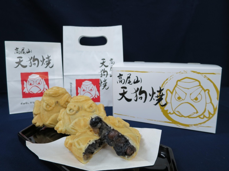天狗の顔をかたどった焼き菓子の天狗焼が黒い皿に並び、手前の1つは割られて黒豆入りの餡が見えている。奥には高尾山天狗焼と書かれた紙袋2つと天狗の絵が入った白い箱がある