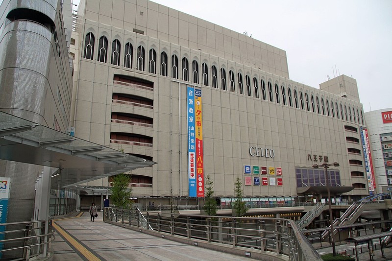曇り空の下のセレオ八王子北館の外観。ベージュの外壁の上部にアーチ形の窓が並び、中央にCELEOと八王子駅の文字、壁に縦長の垂れ幕がある。手前にJR八王子駅北口のペデストリアンデッキが続いている