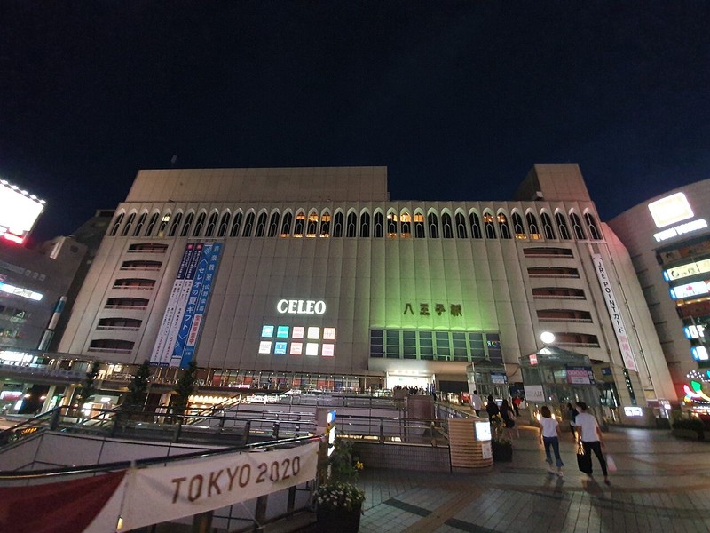夜のセレオ八王子北館の外観。白い外壁の上部にアーチ形の窓が並び、中央にCELEOの白い文字と緑に光る八王子駅の文字がある。手前はJR八王子駅北口のペデストリアンデッキで、通行人が歩いている