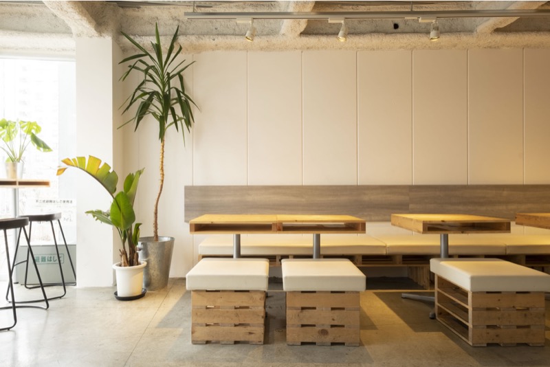 白い壁と木の床の店内。パレットを重ねたテーブルに白いスツールが並び、観葉植物と照明が置かれている